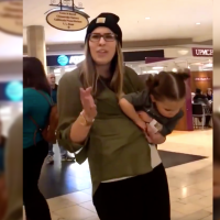 Mother holding her toddler daughter pointing at person filming. She's wearing a black beanie and a green blouse.