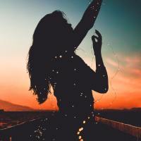 woman silhouetted by sunrise holding blurred fairy lights and hands raised to sky
