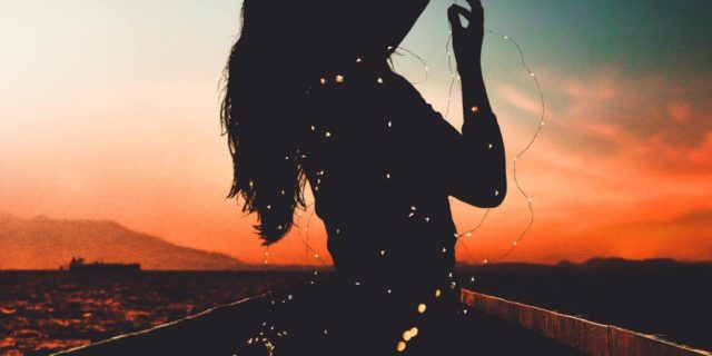 woman silhouetted by sunrise holding blurred fairy lights and hands raised to sky