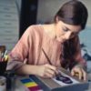 woman designing box or painting on table surrounded by art supplies