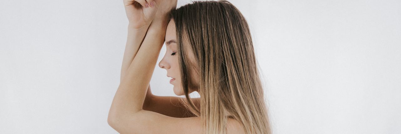 side view of woman with arms raised to her head and eyes closed against white background