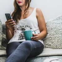 woman looking at phone and drinking coffee while sitting in bed