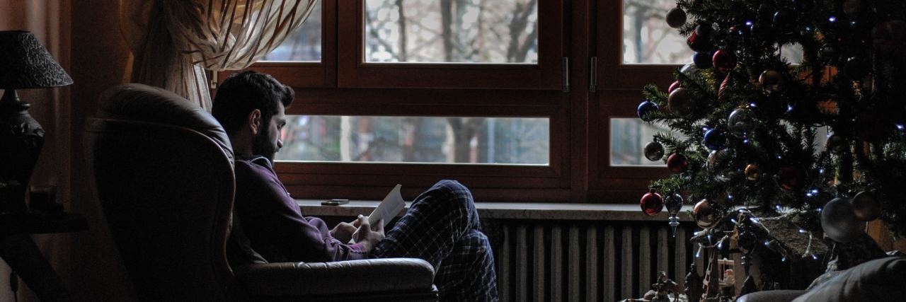 Tips for Challenging the 'Holiday Blues' and Depression This Year man reading book by window at Christmas with tree beside him