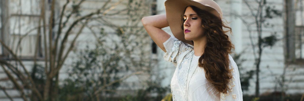 How to Recover From Dependent Abusive Relationship photo of woman standing in front of old house wearing hat over her long brown hair and her eyes are closed, her hand raised to her hair
