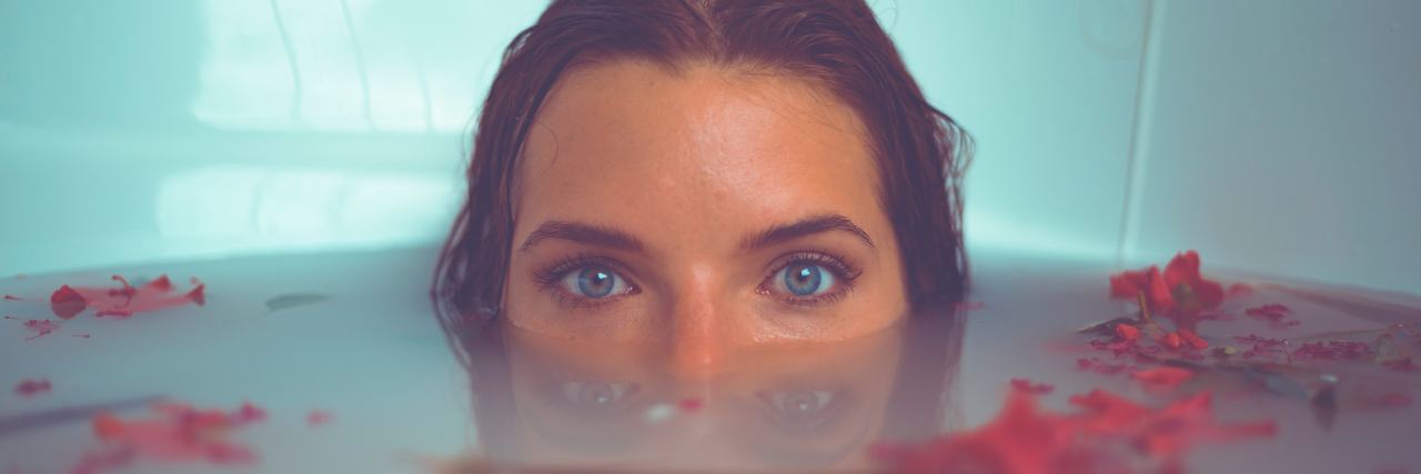 What to Know About Self-Care If You’re Cynical woman partially submerged in bath with flowers looking into camera