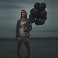 man standing in ocean under stormy sky holding black balloons