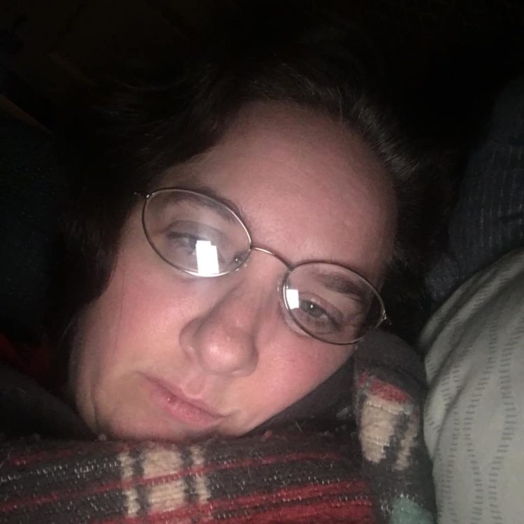 woman lying in bed under a plaid blanket. she's wearing glasses that are slightly askew and her eyes are slightly droopy