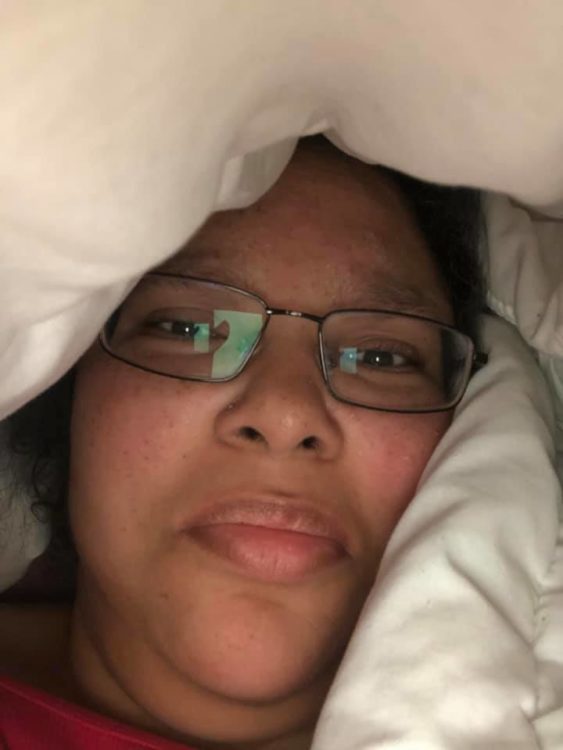 A selfie of a woman with brown skin and glasses, surrounded by a white blanket