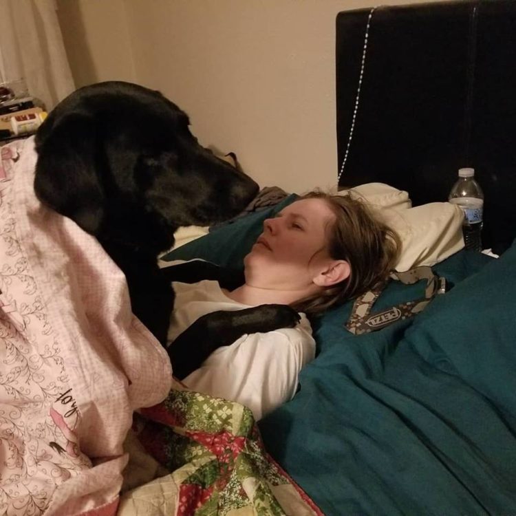 A woman lying in bed. A black dog lies on top of her, his paws rested on her chest.