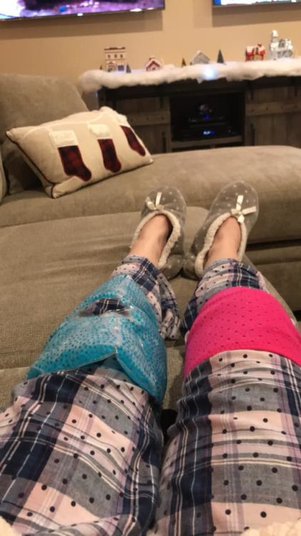 woman sitting on her couch with her legs out straight. she's wearing pajama pants and slippers and has ice packs on her knees