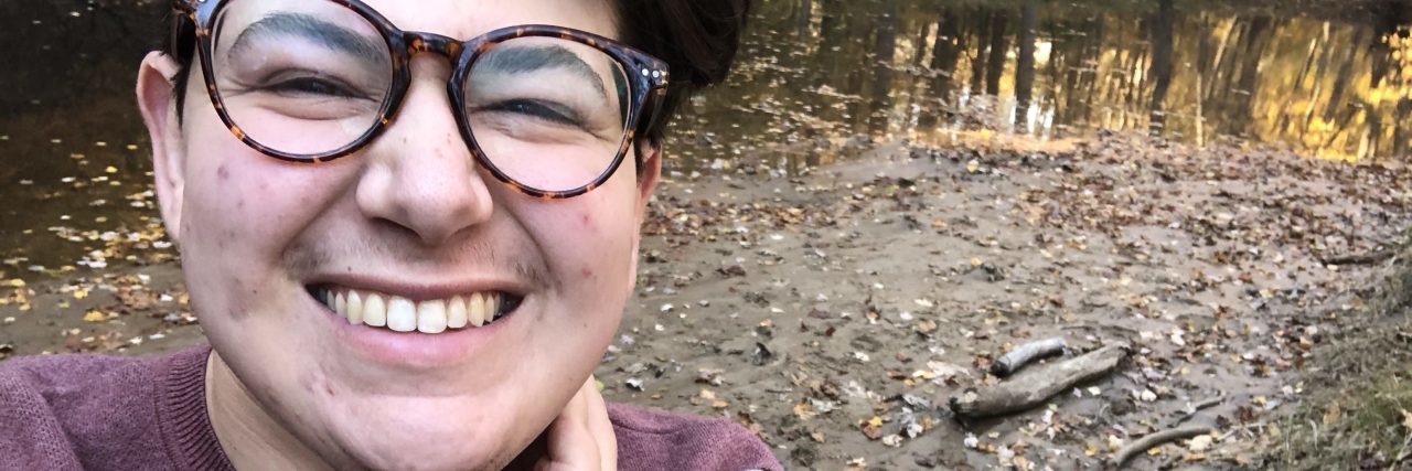 What It's Like to Be Trans and Have Chronic Illness photo of the author standing outside and smiling. they're wearing a red sweater and glasses