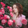 photo of woman standing beside pink roses with eyes closed and hands raised to flowers