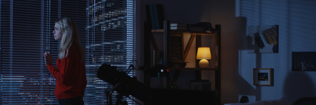 The Shame I Felt Before and After My Brother's Death Medium wide shot of a lonely woman looking out a window at night then walking away