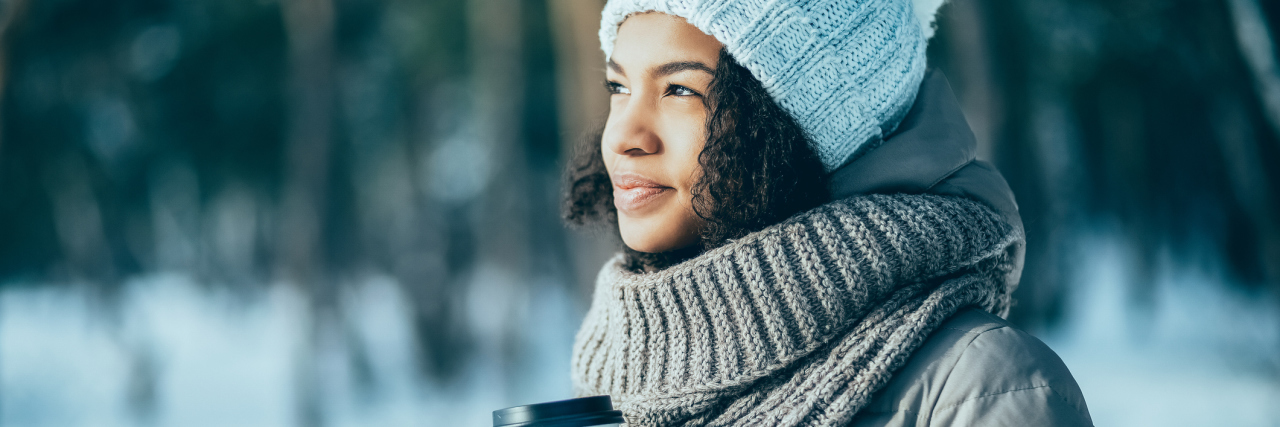 Practicing Self-Compassion in the New Year With Chronic Illness woman wearing a hat, coat, scarf and mittens standing outside in the snow and holding a coffee
