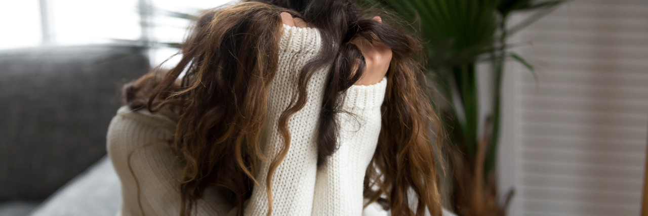 What People Don't Understand About Chronic Migraine woman with long dark hair wearing a white sweater sitting in a chair with her head in her hands
