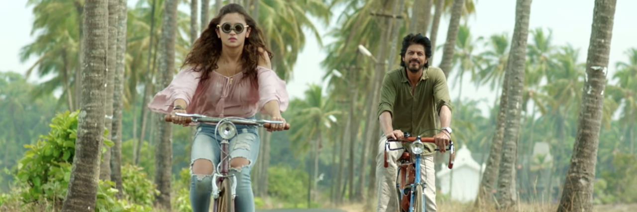 still image of bollywood movie Dear Zindagi showing male and female actors riding down tree-lined road on bicycles