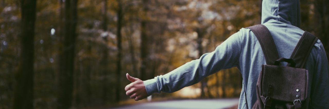 photo of hitchhiker taken from behind with hand and thumb outstretched and autumnal trees