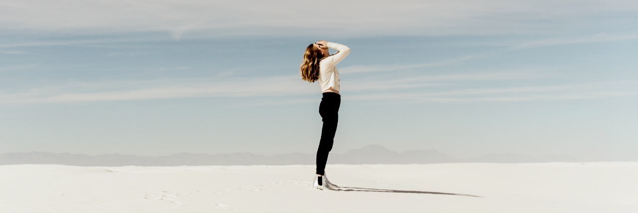 photo of woman standing on white sand or desert, head raises to the cloudy sky with hands covering her face