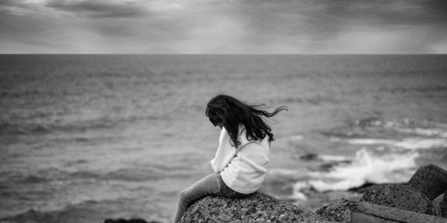 black and white photo of woman sitting on rocks by ocean under stormy sky