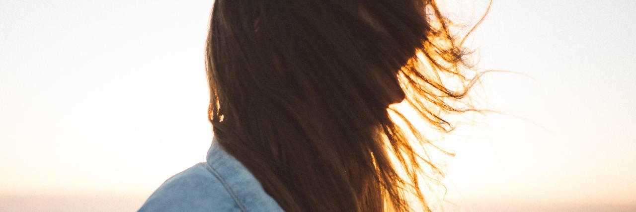 woman wearing a denim jacket and standing on the beach at sunset with her hair hanging in front of her face