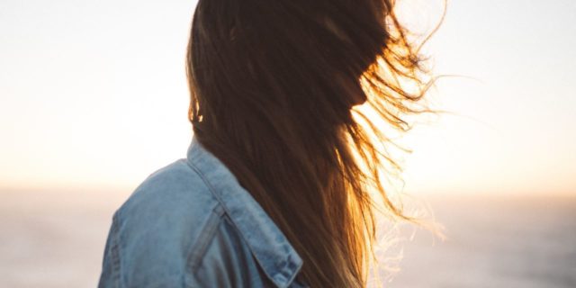 I May Be Chronically Ill, but There Is No Need to Pity Me woman wearing a denim jacket and standing on the beach at sunset with her hair hanging in front of her face