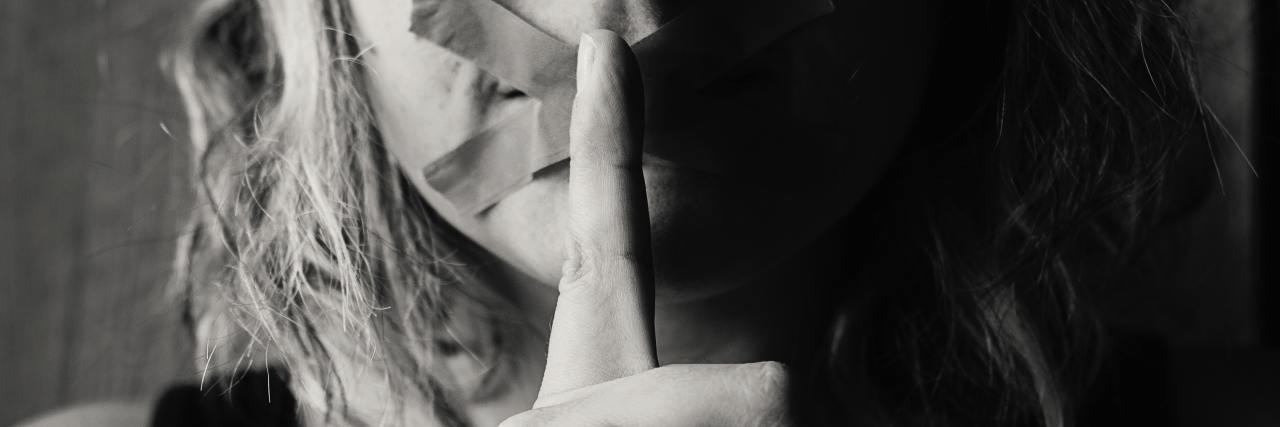 Silence With Selective Mutism Isn't What You Think black and white photo of woman with X tape across mouth and holding finger to mouth in hush gesture