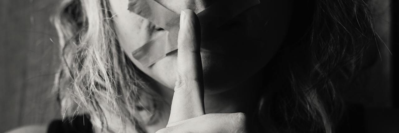 black and white photo of woman with X tape across mouth and holding finger to mouth in hush gesture