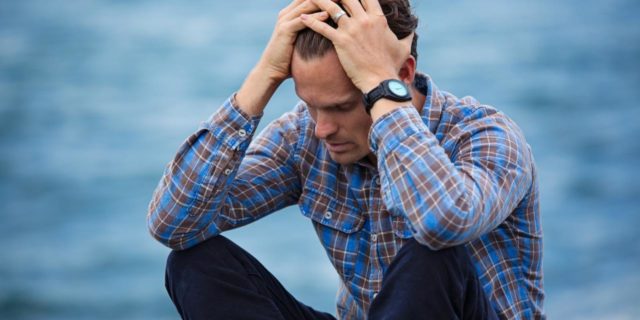 photo of man depressed sitting by ocean with hands on forehead
