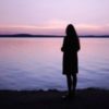 photo of woman silhouetted against ocean or lake at sunset with lilac sky and distant clouds