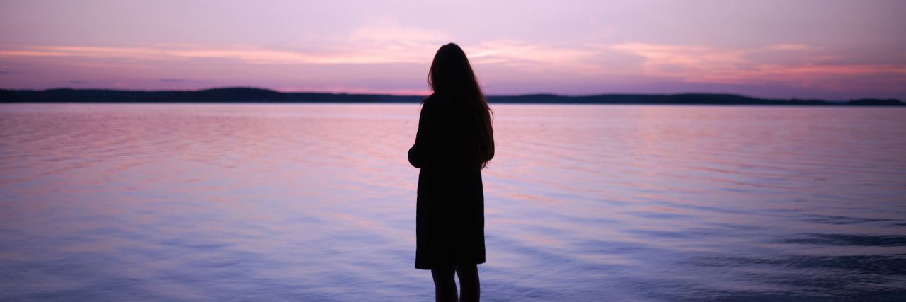 I Feel Attached to My Trauma photo of woman silhouetted against ocean or lake at sunset with lilac sky and distant clouds