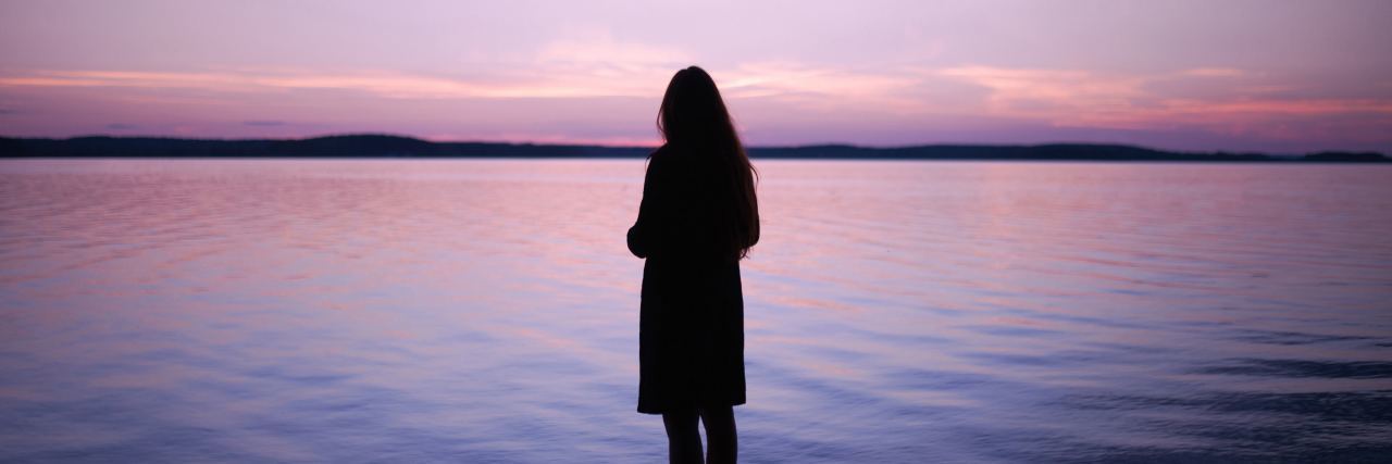 photo of woman silhouetted against ocean or lake at sunset with lilac sky and distant clouds