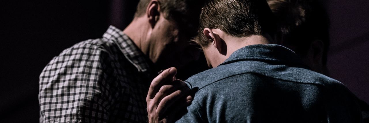 two men in semi darkness with one man's hand on the other's shoulder in support or prayer