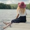 photo of blonde woman sitting on pier at lake looking at water and trees