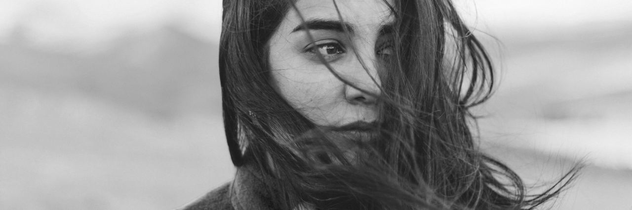 black and white photo of woman with long hair standing outside. she's looking over her shoulder and the wind is blowing several strands of her hair across her face.