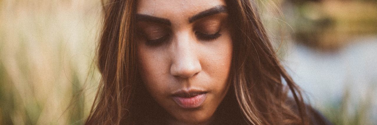 Struggling With Guilt When You Live With Chronic Illness close up photo of a woman with long dark hair looking down and standing in tall grass near a pond