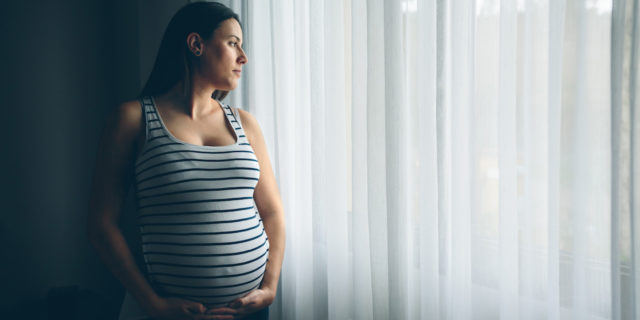A pregnant woman standing by a window covered in white curtains