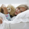 a sick girl is laying in a bed hugging a teddy bear