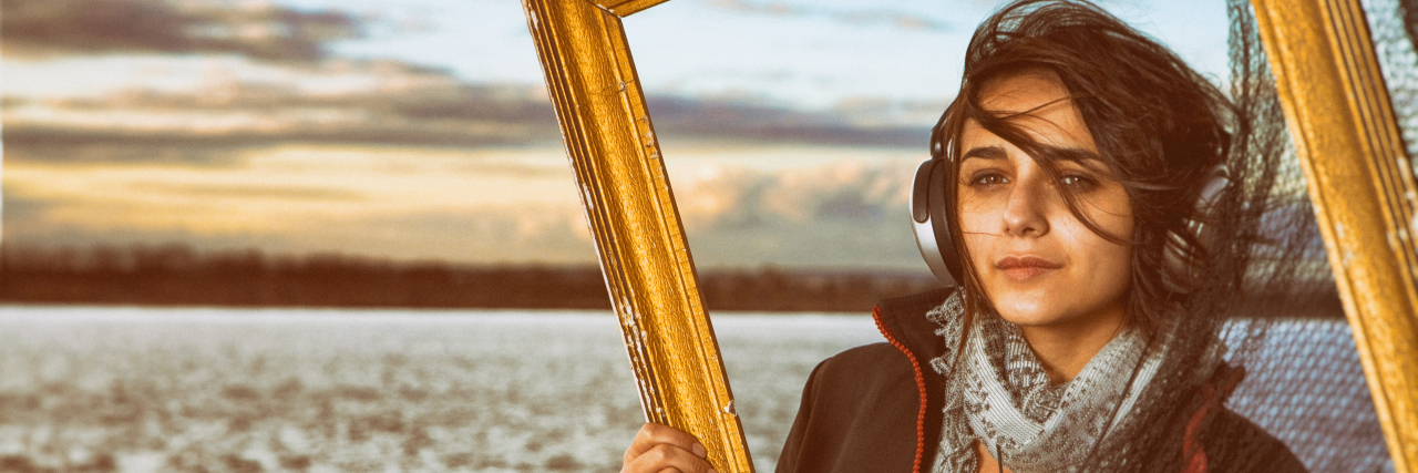 Woman wearing headphones looking through frame, with ocean in the background.