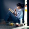 photo of woman sitting by open fridge surrounded by snacks and reading book