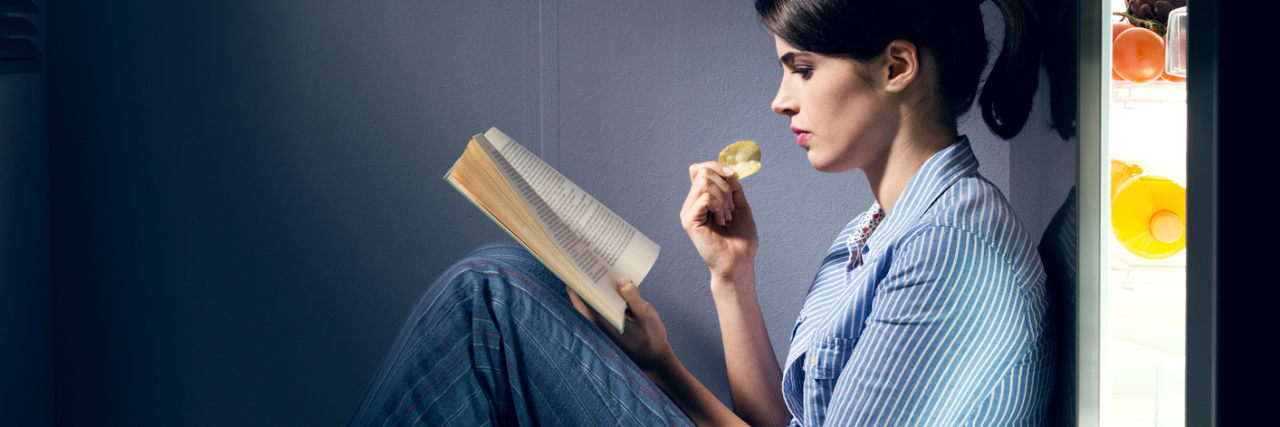 photo of woman sitting by open fridge surrounded by snacks and reading book