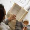 woman sitting in bed reading a book and holding a mug of tea
