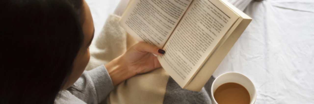 Why Learning Self-Care Is a Process as Someone With Multiple Sclerosis woman sitting in bed reading a book and holding a mug of tea