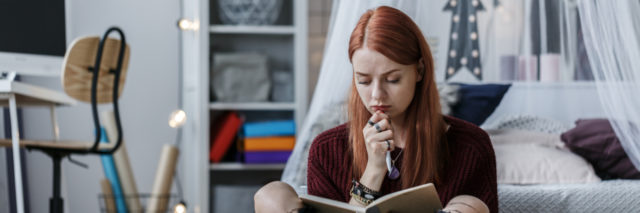 Cogitative, redhead girl thinking over a notebook while sitting on a floor in her room