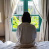 Lone woman sitting on the bed looking out at the window in the morning