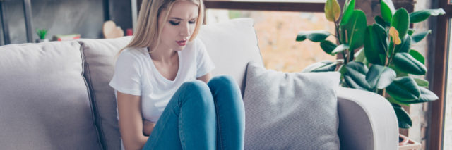 photo of blonde woman sitting on couch looking unwell or anxious with hands across stomach