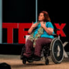 Jamie Smith sitting in her wheelchair on stage in front of TEDx sign, giving a speech.