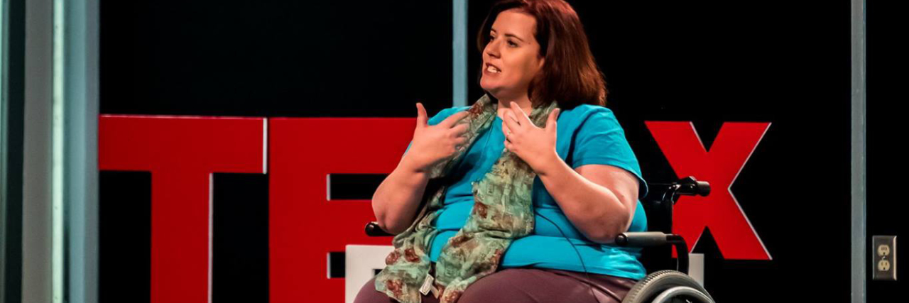 Showing True Empathy When Interacting With People With Disabilities Jamie Smith sitting in her wheelchair on stage in front of TEDx sign, giving a speech.