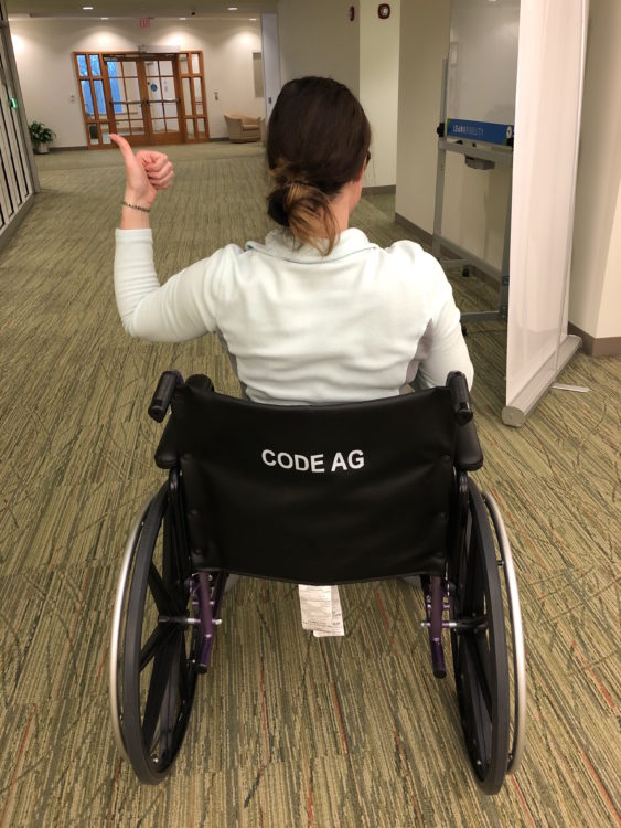 A wheelchair user using a thumb up, facing away. On the back of her wheelchair, it reads CODE AG
