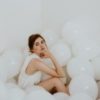 photo of woman wearing white dress sitting in white room surrounded by white balloons, resting chin on her hand