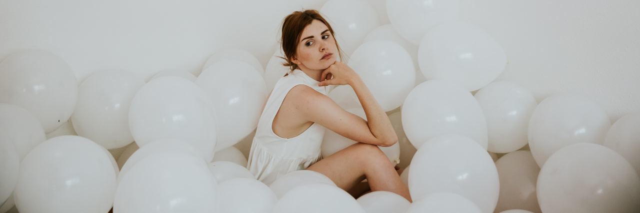 What I Wish Others Knew About Social Anxiety and Being Autistic photo of woman wearing white dress sitting in white room surrounded by white balloons, resting chin on her hand